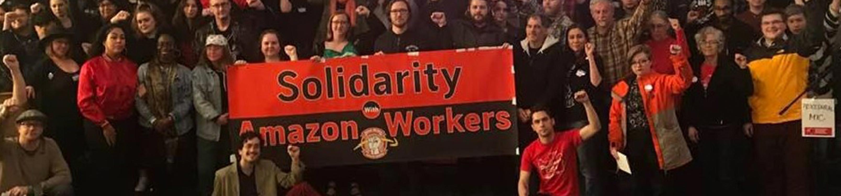 Image of a crowd with raised fists standing behind an Solidarity with Amazon Workers" banner.
