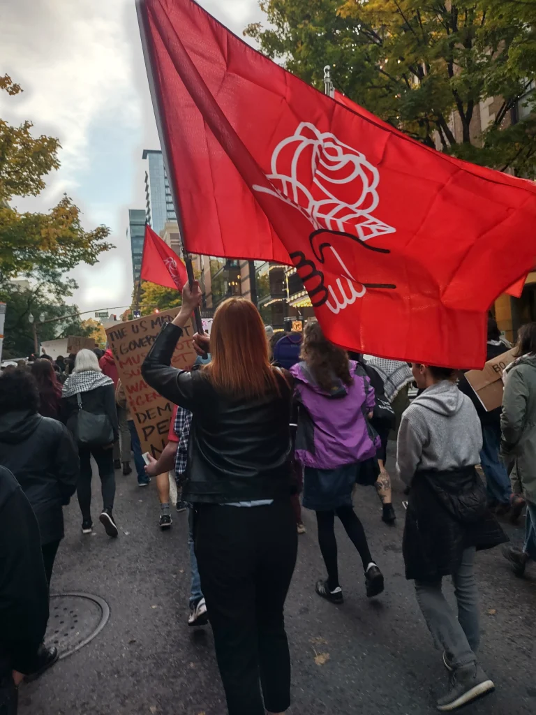 Demonstrators marching in downtown Portland holding cardboard signs and hoisting large DSA flags.